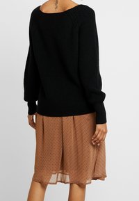 Žena v černém žebrovaném svetru přes hnědou sukní po kolena s malým černým tečkovaným vzorem, stojící na jednobarevném pozadí.