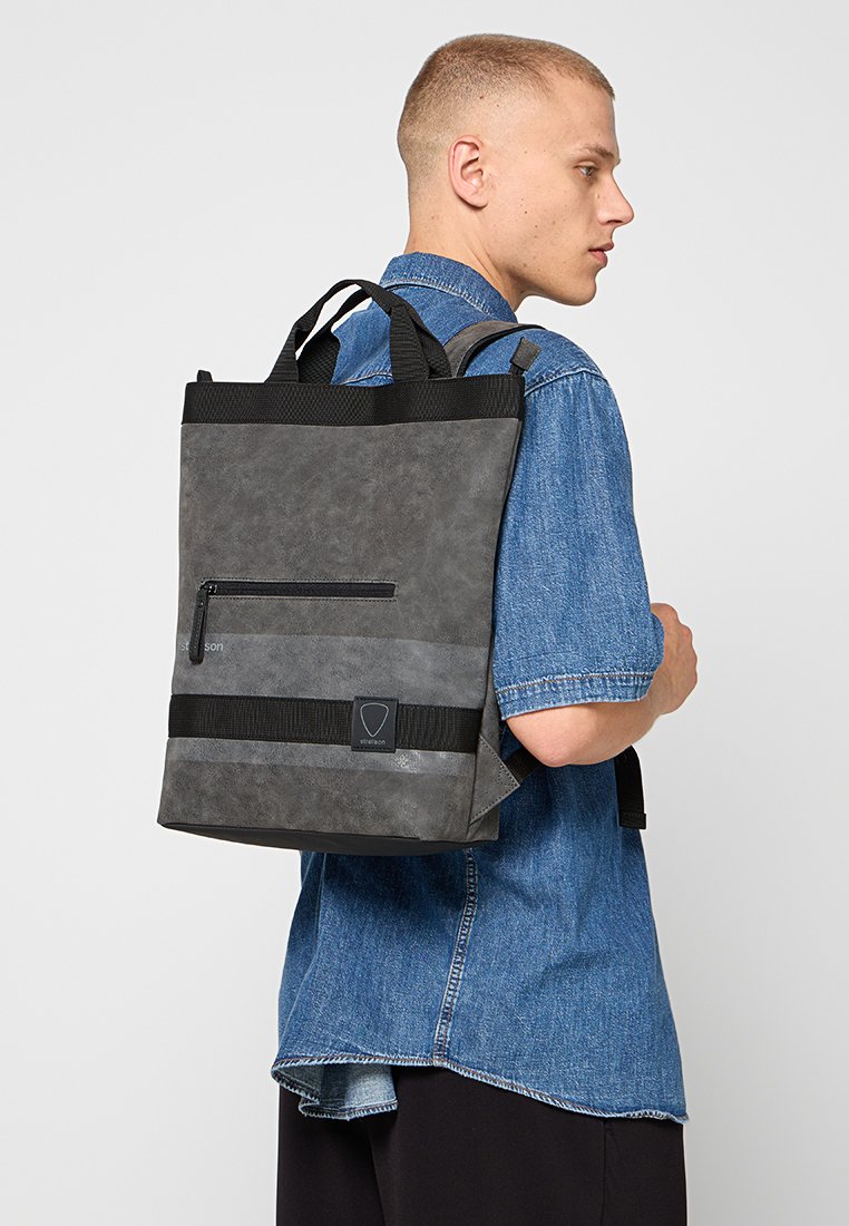 Zaino grigio con finitura testurizzata, dotato di una tasca con zip frontale, spallacci neri larghi e un patch con logo. Design strutturato con forma rettangolare.