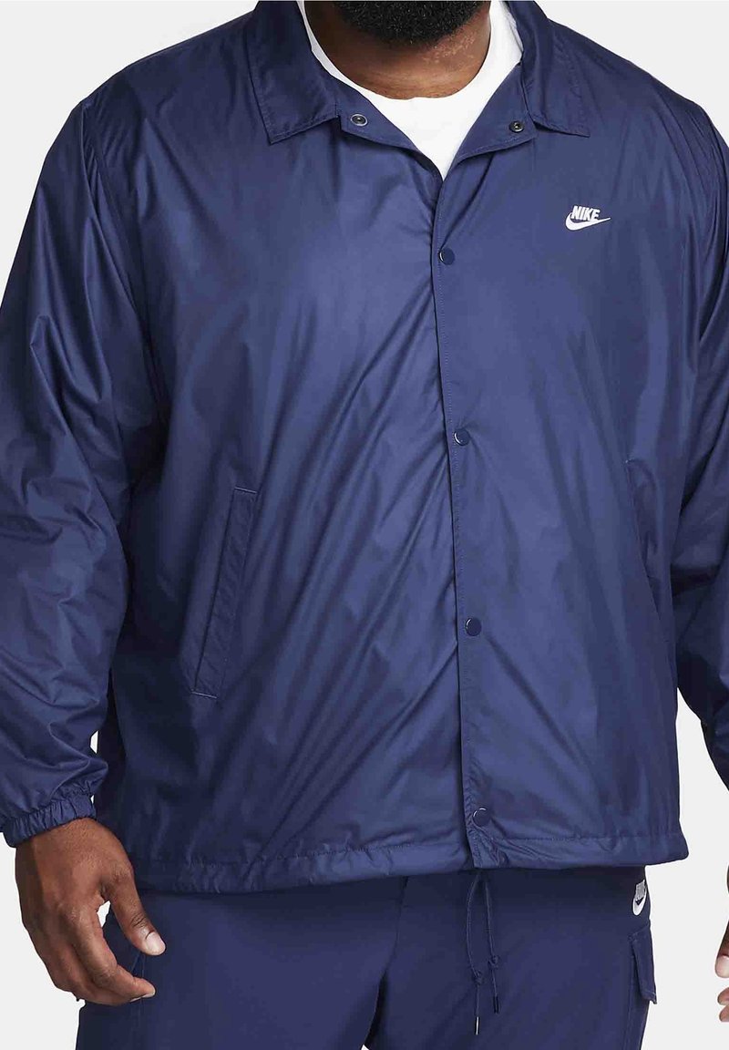 Marinblå nylonjacka med tryckknappsstängning, elastiska ärmslut och en sidficka på vänster sida; har en liten vit Nike-logotyp på bröstet.