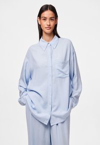 Camisa de botones de manga larga azul claro con cuello clásico y un bolsillo en el pecho, con un corte relajado y una textura de tejido suave.