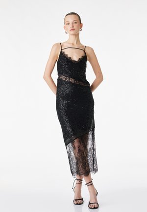 Femme portant une robe noire à sequins avec des détails en dentelle, des bretelles spaghetti et un ourlet asymétrique en dentelle transparente, associée à des talons noirs à brides.