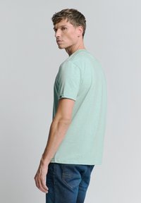 Lichtgroene T-shirt met korte mouwen in een relaxed fit, met subtiele textuur en een klein logo-accent op de mouw, gecombineerd met donkerblauwe spijkerbroek.