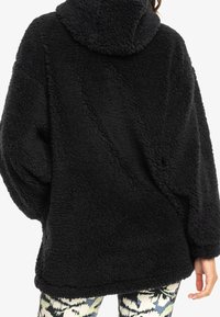 Zwarte fleece pullover met capuchon, voorzien van een zachte, gestructureerde uitstraling, oversized pasvorm en verlaagde schouders. Geen zichtbare sluitingen.
