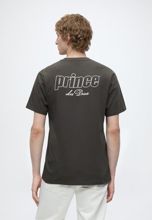 Ung person med krøllet blondt hår iført mørkegrå t-shirt med teksten "prince Les Deux" på ryggen, kombineret med hvide bukser, stående mod en ensfarvet baggrund.