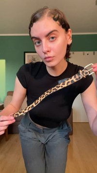 Pas za leopardji vzorec z teksturiranim površjem, z srebrno zaponko in ozko obliko, ga drži oseba, oblečena v črno majico.