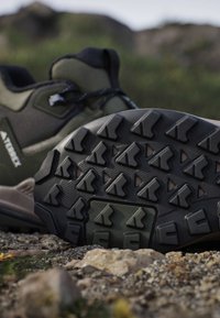 Hiking boot lying on rocky ground showing detailed tread pattern on the sole with outdoor scenery blurred in background.