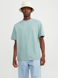 T-shirt en coton bleu clair avec des manches courtes, une coupe décontractée et un logo brodé sur la poitrine. Porté avec un jean bleu clair et un chapeau seau beige.