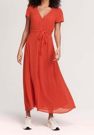 Femme portant une robe maxi rouge fluide à manches courtes boutonnée avec une ceinture à la taille, associée à des baskets blanches, debout sur un fond uni.