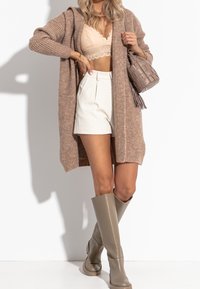 Beige gestrickte Strickjacke, Spitzen-Bralette, weiße High-Waist-Shorts und beige kniehohe Stiefel. Außerdem enthält es eine strukturierte beige Tasche.