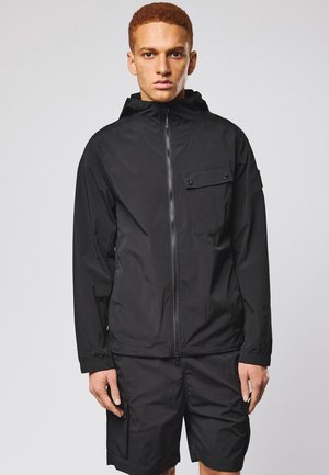 Młody mężczyzna o krótkich rudych włosach, ubrany w czarną bluzę z kapturem na zamek oraz pasujące czarne szorty, stoi na tle jednolitego tła.