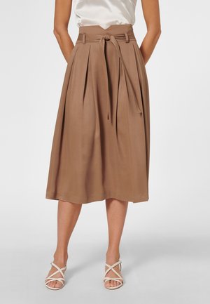 Femme portant une jupe midi taille haute couleur beige avec une ceinture nouée, associée à des sandales blanches à brides et un top blanc sans manches.