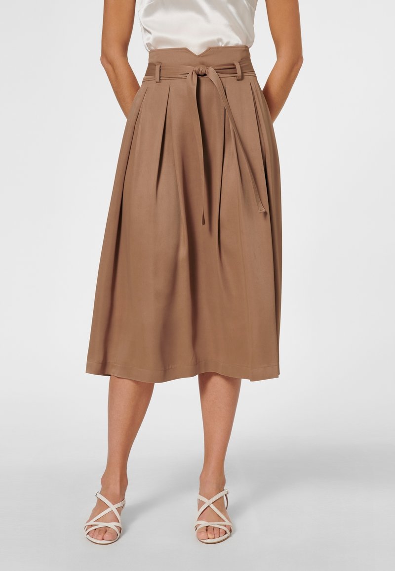 Femme portant une jupe midi taille haute couleur beige avec une ceinture nouée, associée à des sandales blanches à brides et un top blanc sans manches.
