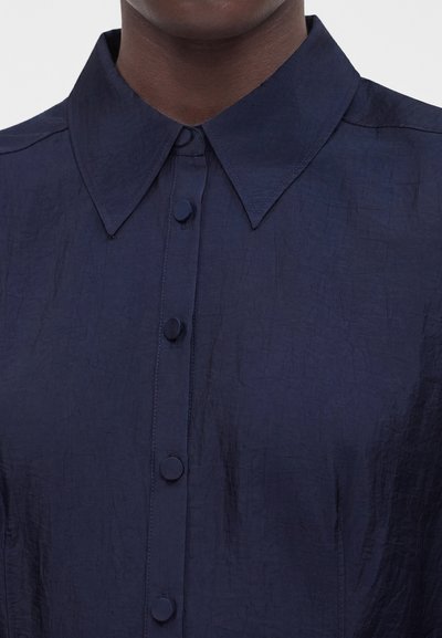 Άνδρας που φοράει σκουρόχρωμο μπλε πουκάμισο με κουμπιά, με φαρδιά, μυτερή γιακά και κουμπιά καλυμμένα με ύφασμα.