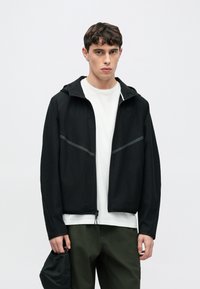 Czarna kurtka na zamek błyskawiczny z kapturem, wykonana z gładkiego materiału. Posiada odblaskowe elementy i jest zestawiona z białą koszulą oraz zielonymi spodniami.