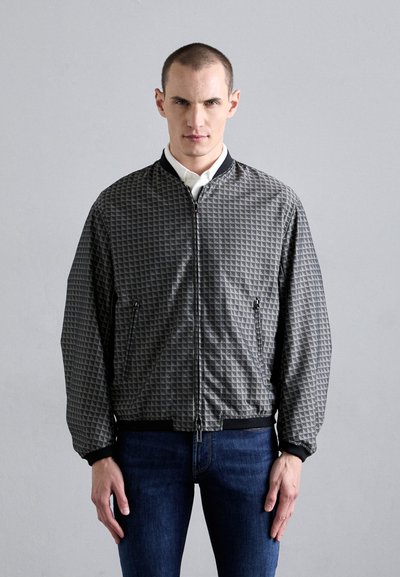 Giacca bomber grigia con motivo, superficie testurizzata, chiusura a zip, polsini e orlo a coste. Indossata sopra una camicia bianca con colletto e jeans blu.