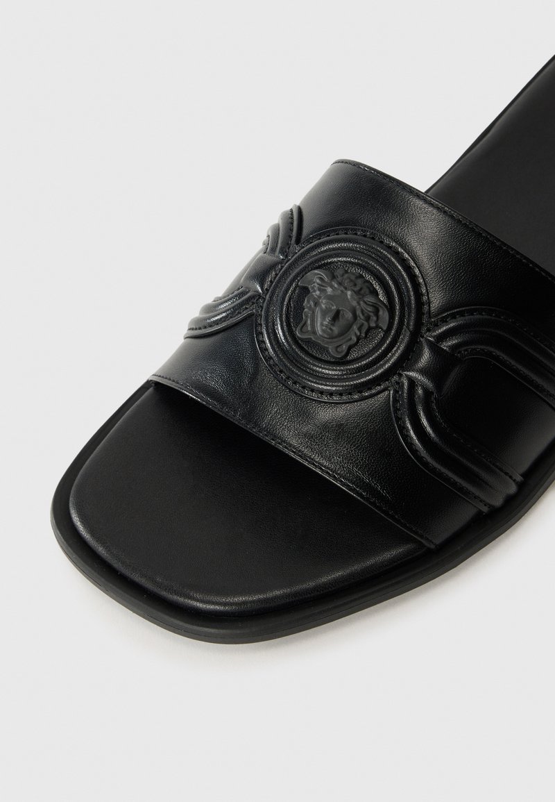 Schwarze Leder-Sandale mit breitem Riemen, der ein geprägtes Löwenkopf-Emblem und geschwungene Details aufweist, flacher Sohle und matter Oberfläche.