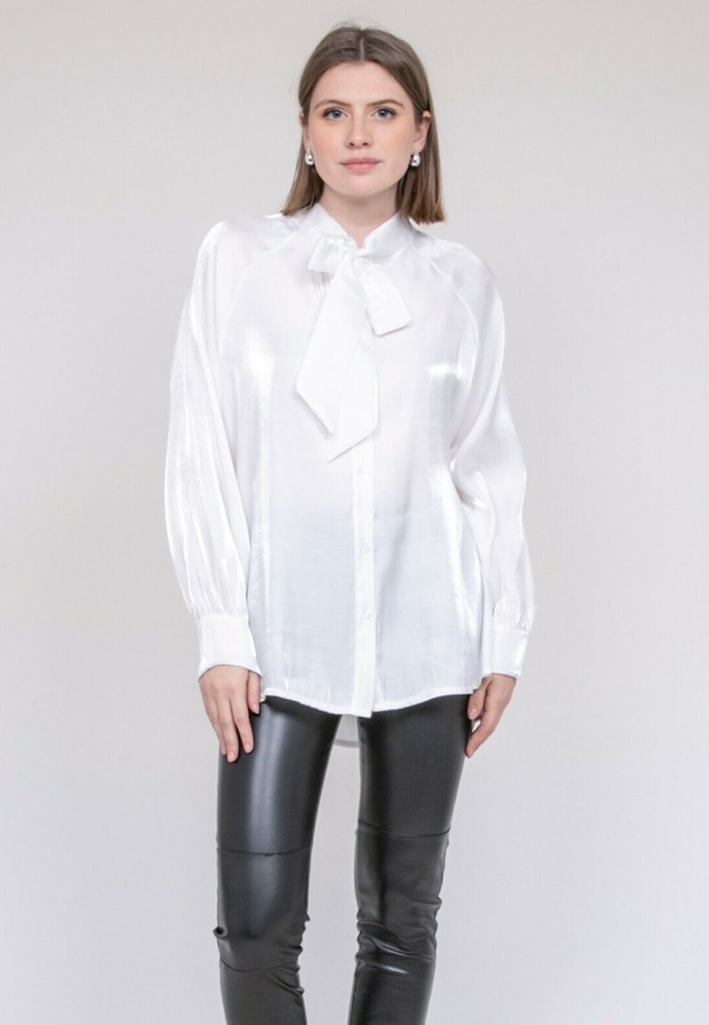 Blouse en satin blanc à manches longues avec un nœud noué au col. Associée à un pantalon en cuir noir ajusté. Texture lisse et brillante.