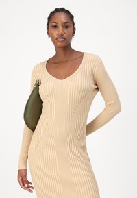 Abito lavorato a rete beige con maniche lunghe e scollatura a V, abbinato a una borsa a tracolla di forma ovale verde con accenti in metallo dorato.