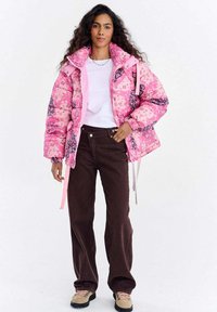 Różowa kurtka puchowa z kwiatowymi i geometrycznymi wzorami, zestawiona z ciemnobrązowymi szerokimi spodniami. Beżowe buty z różowymi sznurowadłami dopełniają strój.