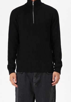 Homme portant un pull noir côtelé avec col à demi-fermeture éclair et un jean gris foncé, debout devant un fond blanc.