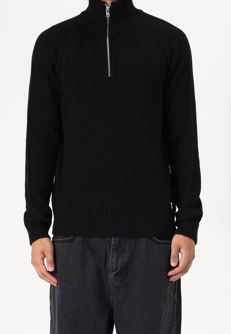 Homme portant un pull noir côtelé avec col à demi-fermeture éclair et un jean gris foncé, debout devant un fond blanc.