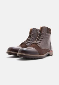 Botas de tobillo de cuero marrón y ante con punta redondeada, cordones en la parte delantera y cremallera, que presentan una suela texturada y detalles en rojo.