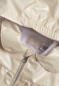 Veste imperméable beige clair avec texture brillante, motif à pois, fermeture éclair et poignets élastiques. Étiquette visible à l'intérieur du col.