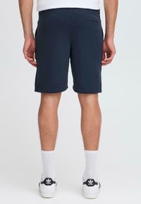 Homme portant un short bleu marine, des chaussettes blanches et des baskets Adidas blanches, se tenant de dos face à la caméra sur un fond uni.