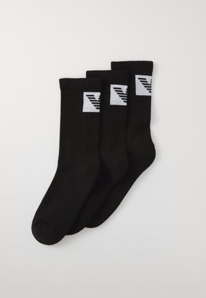 Trois paires de chaussettes en coton noir avec une texture côtelée. Chaque chaussette est dotée d'une étiquette de logo blanche avec des rayures diagonales noires.