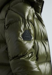 Olive zaļā pūkainā jaka ar spīdīgu tekstūru. Funkcijas ietver melnu gumijas logotipa uzšuvumu uz augšdelma un quilted daļas siltumizolācijai.