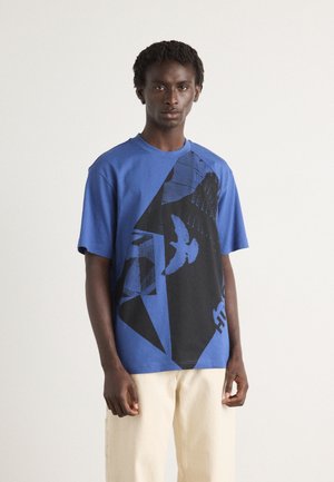 Blå bomulds T-shirt med et stort sort abstrakt grafisk og fugledesign foran. Korte ærmer, rund hals, afslappet pasform.