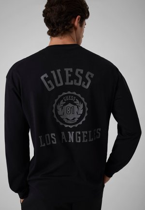 Uomo che indossa una maglietta nera a maniche lunghe con "GUESS LOS ANGELES" e "81" stampati sulla schiena in lettere grigio scuro.