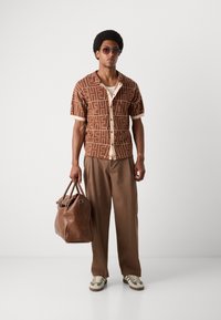 Short-sleeve brown shirt with geometric pattern, beige accents, paired with loose brown trousers and a brown leather duffel bag.