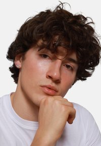 Gros plan sur un jeune homme aux cheveux bruns bouclés, portant un t-shirt blanc, mettant en valeur une boucle d'oreille en argent et une peau légèrement bronzée.