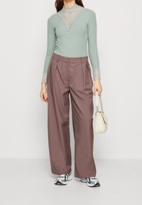 Mátově zelený svetr s dlouhým rukávem a krajkovými detaily na výstřihu, spárovaný s hnědými kalhotami s vysokým pasem a širokými nohavicemi a bílou kabelkou. Texturovaná tkanina.