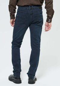 Jean slim fit bleu foncé avec une texture lisse, présentant deux poches arrière, des coutures décoratives et une taille avec des passants de ceinture. Chaussures noires.