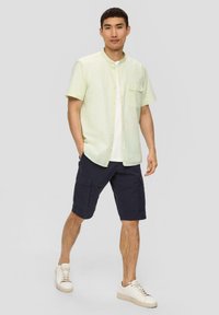 Lichtgele kortarm knoopshirt met een borstzak, gecombineerd met donkerblauwe cargoshorts en witte sneakers.