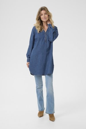 Kvinde i en lang blå denimskjorte, lyseblå jeans og brune ankelstøvler, der står med den ene hånd, der rører ved hendes nakke.