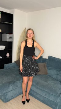 Smilende kvinne i svart ermeløs topp og blomstrete skjørt står ved siden av en blå sofa og et surfebrett i en stue.