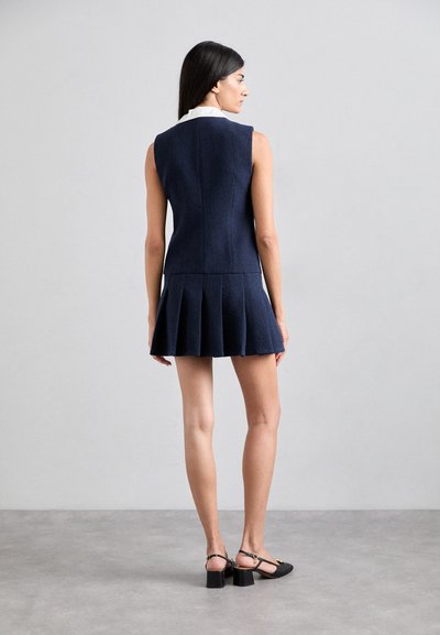 Robe sans manches navy avec une jupe plissée, tissu texturé et un col blanc. Assortie à des chaussures à talons ouverts noires, mettant en valeur un design ajusté.