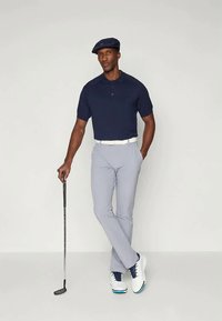 Ναυτική πόλο μπλούζα, ανοιχτό γκρίζες προσαρμοσμένες παντελόνες, λευκή ζώνη και λευκά παπούτσια γκολφ με ναυτικές λεπτομέρειες. Κρατώντας ένα μπαστούνι γκολφ, στέκεται σε απλό φόντο.