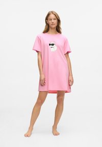 Femeie îmbrăcată cu o rochie lejeră roz, cu un desen al unui câine alb de desene animate purtând ochelari de soare pe față, stând desculță pe un fundal alb.