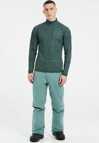 Grünes Zip- up Longsleeve-Top mit hohem Kragen, kombiniert mit hellblauen Hosen. Dunkles Schuhwerk mit strukturiierter Sohle. Einfaches und funktionales Design.