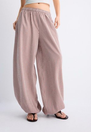 Osoba oblečená v voľných, širokých nohaviciach so vzorom červených a bielych kociek, s elastickým pásom a čiernymi žabkami.