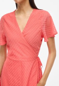 Korálové obalové šaty s krátkymi volánovými rukávmi, s texturovaným zigzagovým vzorom, hlbokým V-výstrihom a viazaním v oblasti pásu.