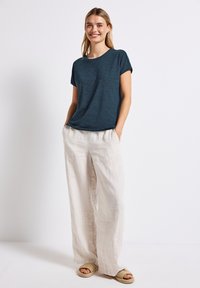 Teal tekstureret kortærmet top, afslappet pasform, parret med løse beige linnedbukser og flettede sandaler, mod en simpel hvid baggrund.