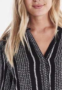 Musta nappikauluspaita, jossa on pystysuoria valkoisia viivoja lehtimäisillä muodoilla ja hienovaraisella tekstuurilla, täydellinen arkikäyttöön.