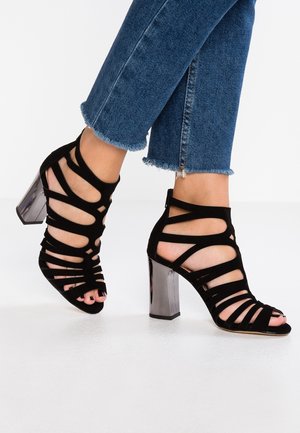 Sorte strop sandaler med høje hæle i suede finish og en skinnende metallisk blokhæle. Åben tå-design med udskæringsdetaljer.