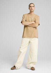 Beige bomulds T-shirt med korte ærmer og et grafisk design på brystet, parret med løstsiddende cremefarvede bukser og brune sko.