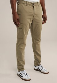 Khaki színű, slim fit cargo nadrág, oldalsó zsebekkel, övbújtatókkal és cipzárral. Fehér sportcipővel viselve, fekete csíkokkal.